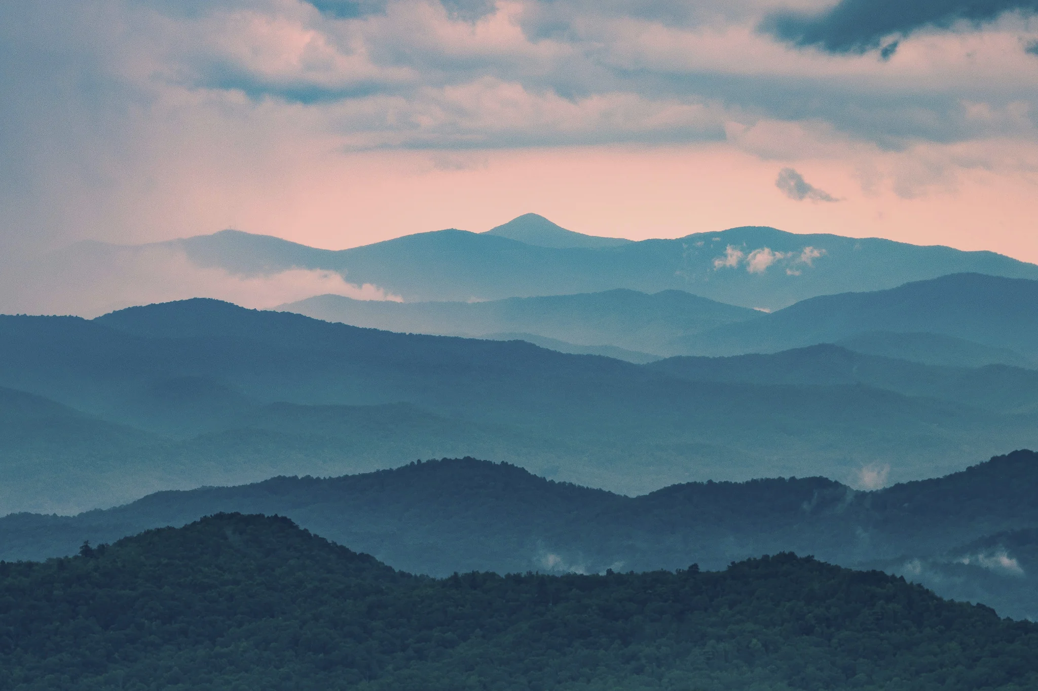 Clouds drifting between layers of forested mountain ridges beneath a soft pink sky.